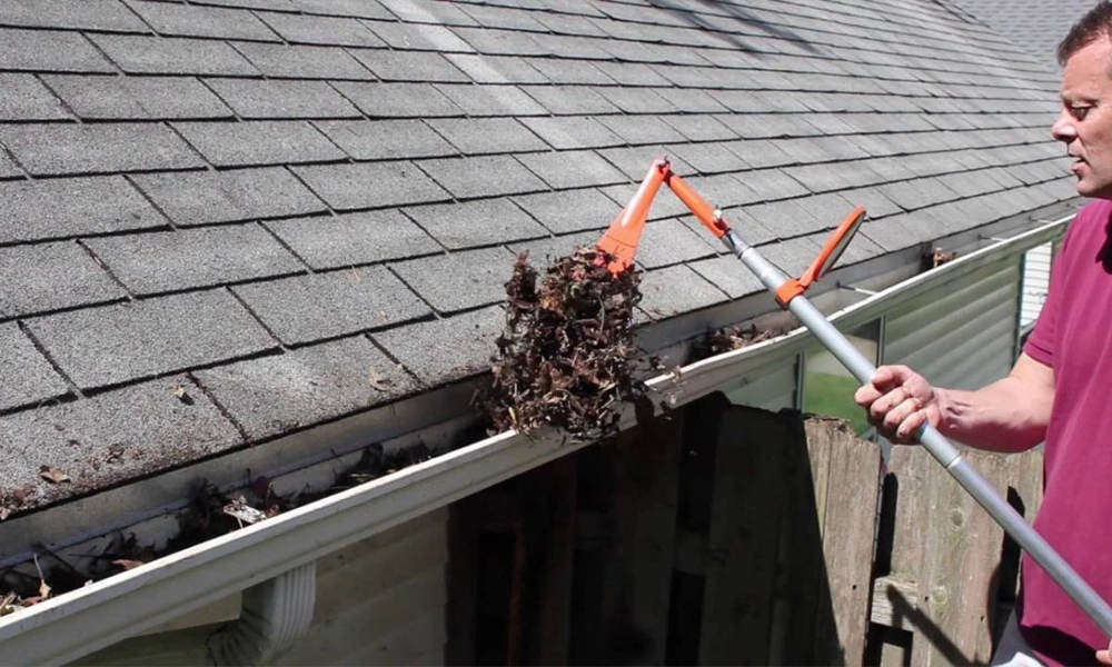 Man using a telescopic gutter cleaning tool to scoop out leaves and debris from a roof gutter."