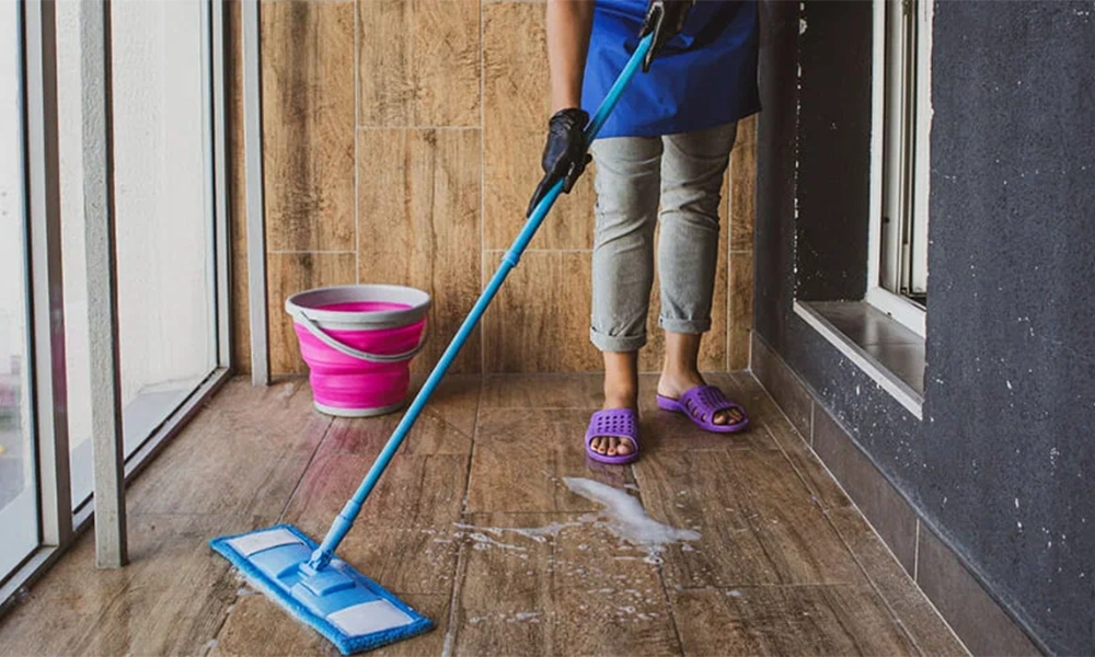 A person cleaning the floor of a balcony with a mop, wearing purple slippers and gloves, with a pink bucket beside them.