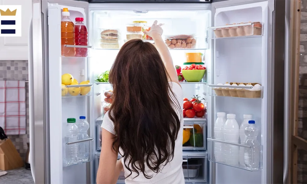 A person reaching into a well-organized refrigerator filled with various food items, such as beverages, fruits, and vegetables.