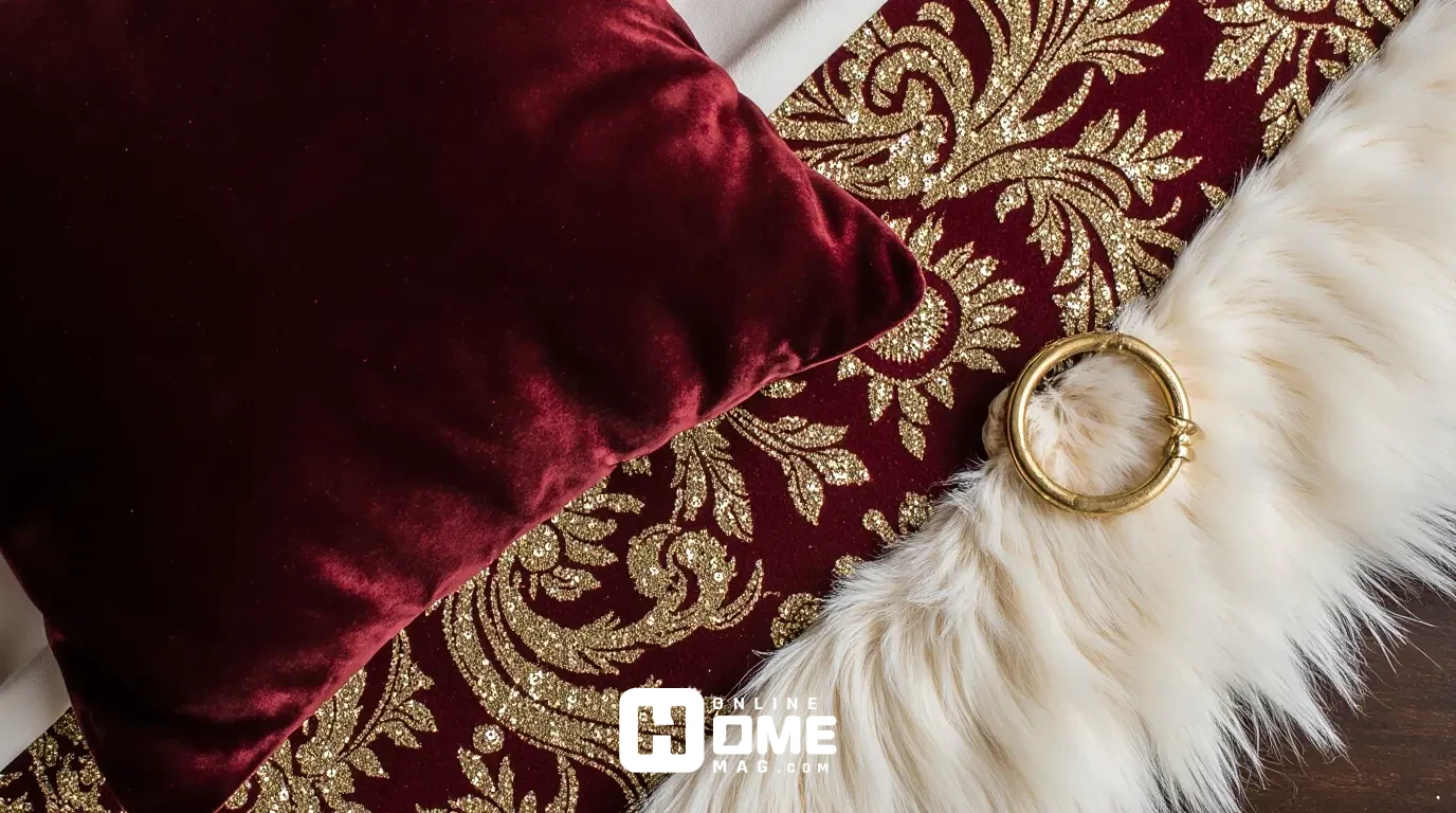 Close-up of layered luxury Valentine's textiles including deep red velvet cushion, gold sequined table runner, burgundy damask napkin with gold napkin ring, and cream faux fur texture, all arranged on a dark wood surface